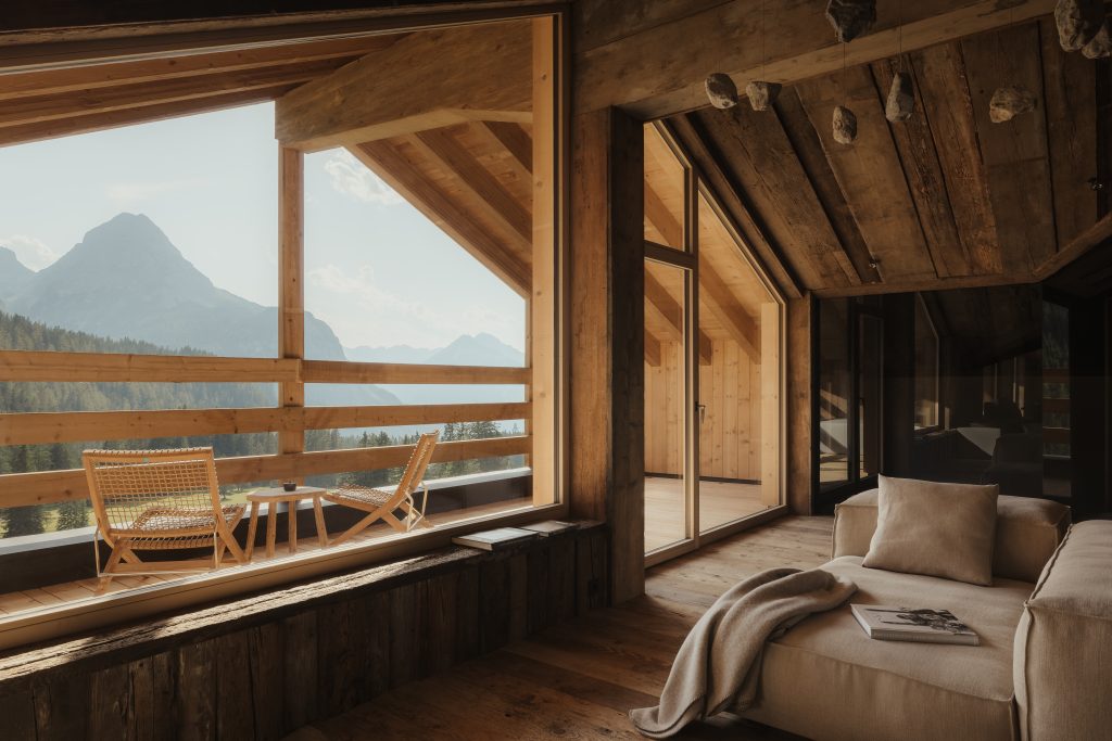 sofa inside a wood room looking out on a terrace with chairs and a table and a view of the Alps in the background