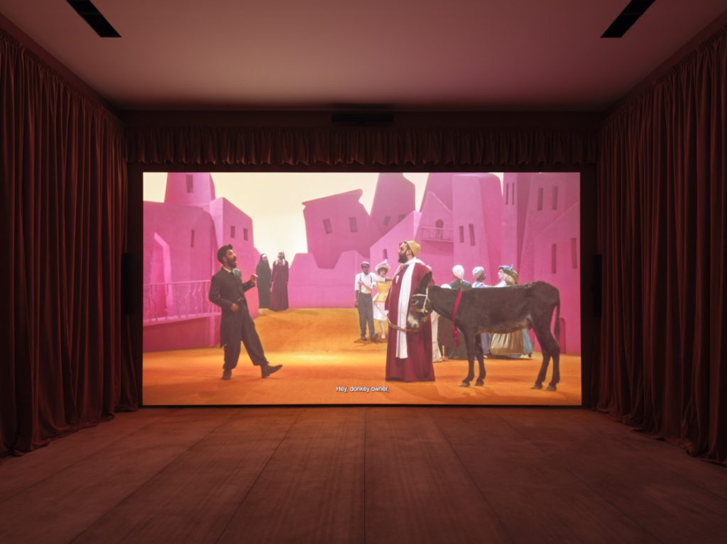 Wael Shawky's Drama 1882 installation shows a video with several people and a donkey playing on a screen in a room with red curtains