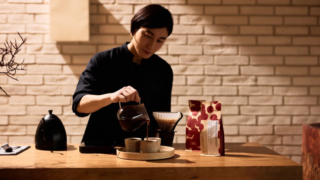 A woman pours coffee into cups on a wood counter, there are bags of coffee and a pour over set