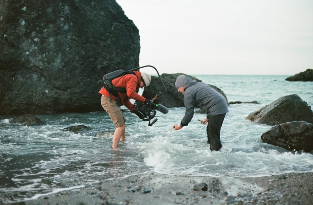 camera person and chef standing ankle-deep in costal waters filming something in chef's hand